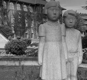 A quietly dramatic sculpture of a boy and a girl is placed outside the St. Eugene Residential School main entrance. A brother and sister, they will soon separated. They touch each other for solace and security, and the girl clenches her fist , clutching her culture, but also perhaps expressing anger and resistance. Photo by Peter Frood